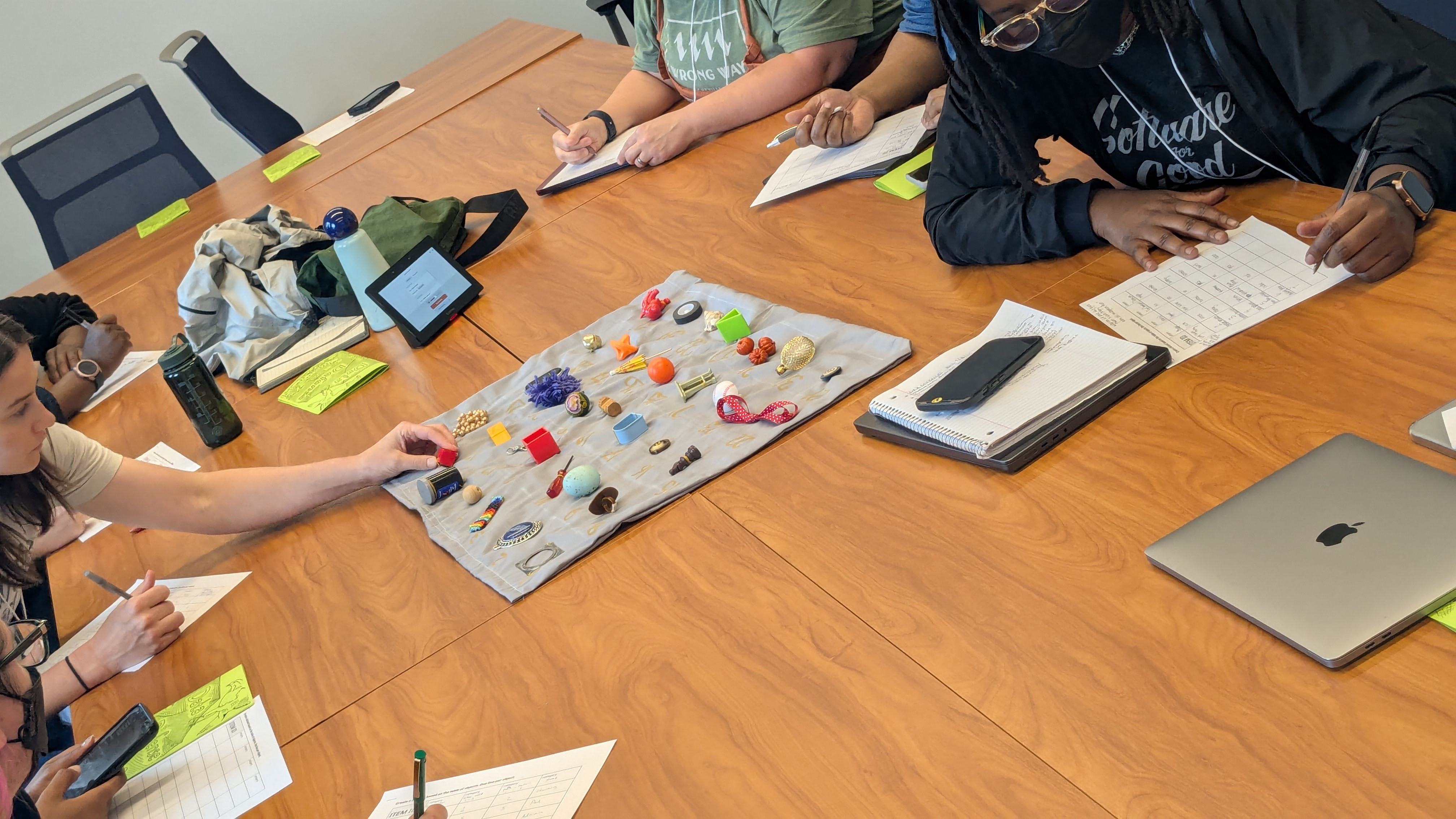 Photo of a wooden table in a conference room where 6 people are seen gathered around with hands reaching out toward a piece of cloth in the middle of the table that has objects embroidered into it representing analog data.